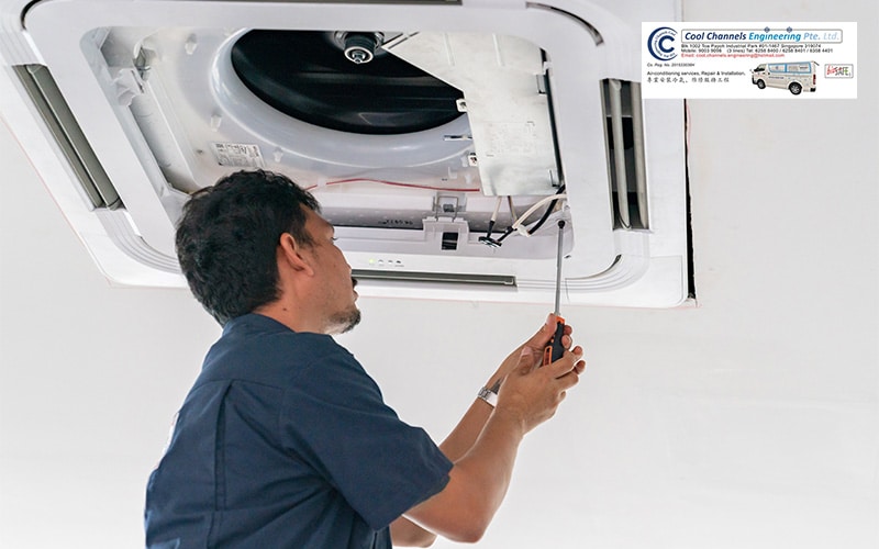 Technician in dark uniform performing maintenance on a ceiling-mounted aircon unit using a screwdriver. Internal components like wiring and fan are visible.