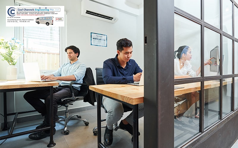 Three people working in a bright office with desks, laptops, glass partition, and wall-mounted aircon units.