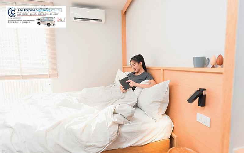 Person sitting up in a neatly made bed with white bedding, reading a book or tablet in a minimalist bedroom featuring light wood furniture, wall-mounted air conditioner, and soft natural light.